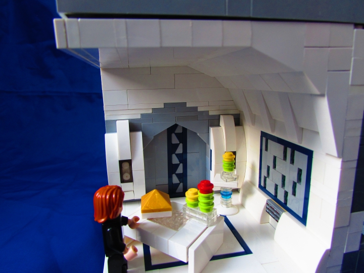 LEGO MOC - New Year's Brick 3015 - Земля. Новый 3015 год.: Один из входов ведет в роскошную с высоким потолком и слепящими белизной стенами. По встроенному в стену серому экрану плавно опускаются голубые снежинки. На изящной мебели установлены символы Нового года - необычные растения с яркими цветами венерианские елки, завезенные туда с Земли и приобретшие новые очертания. Они стали очень популярны в современном обществе, полностью вытеснив обычные земные ели из новогодних жилищ. На столе находится сияющая золотом пирамида, Как символ восхождения к высшей форме сущности человека.